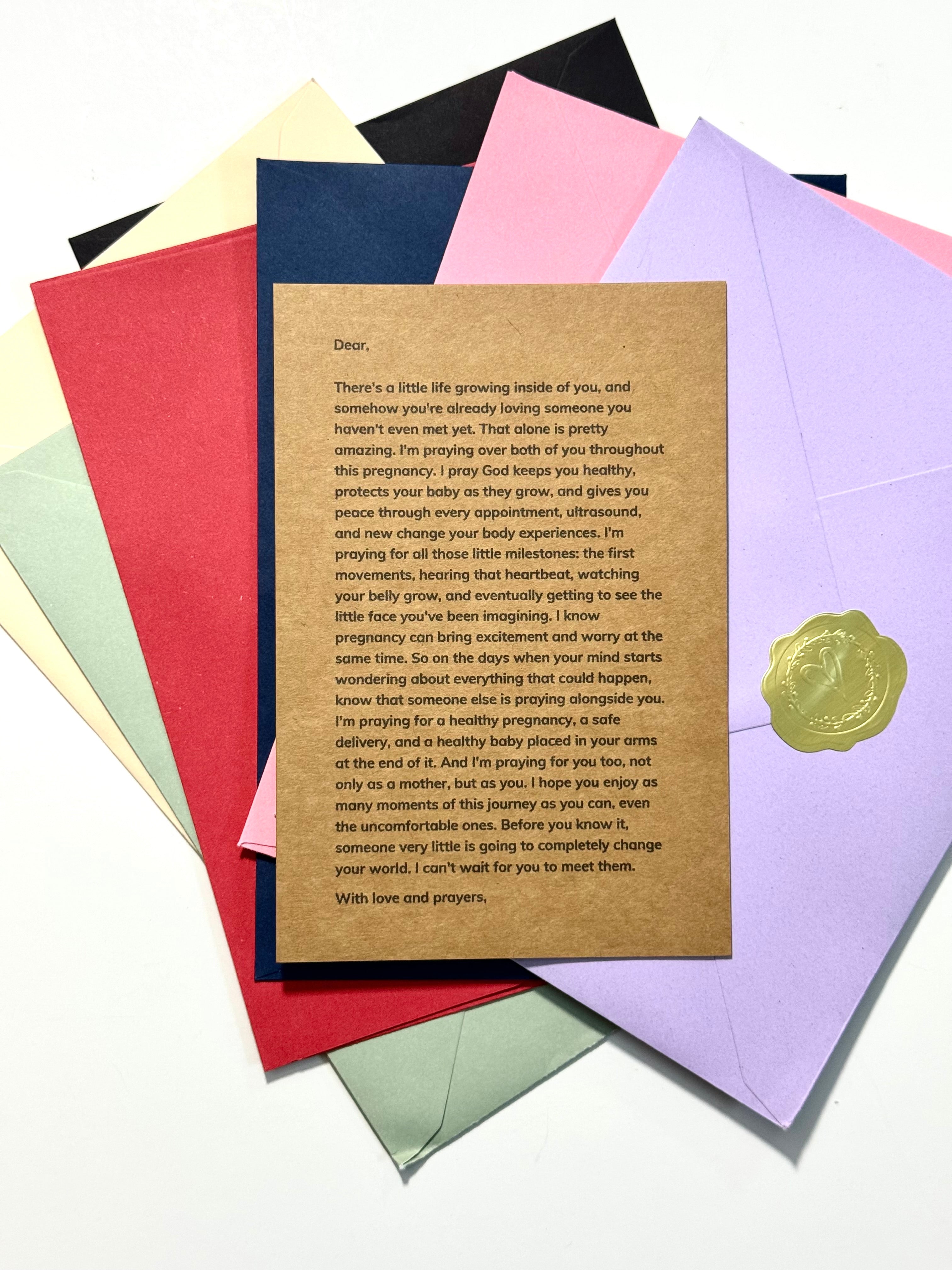 A printed baby shower card with a brown kraft-paper style center and black text, placed on top of several colorful envelopes in red, yellow, blue, pink, and purple on a white background.