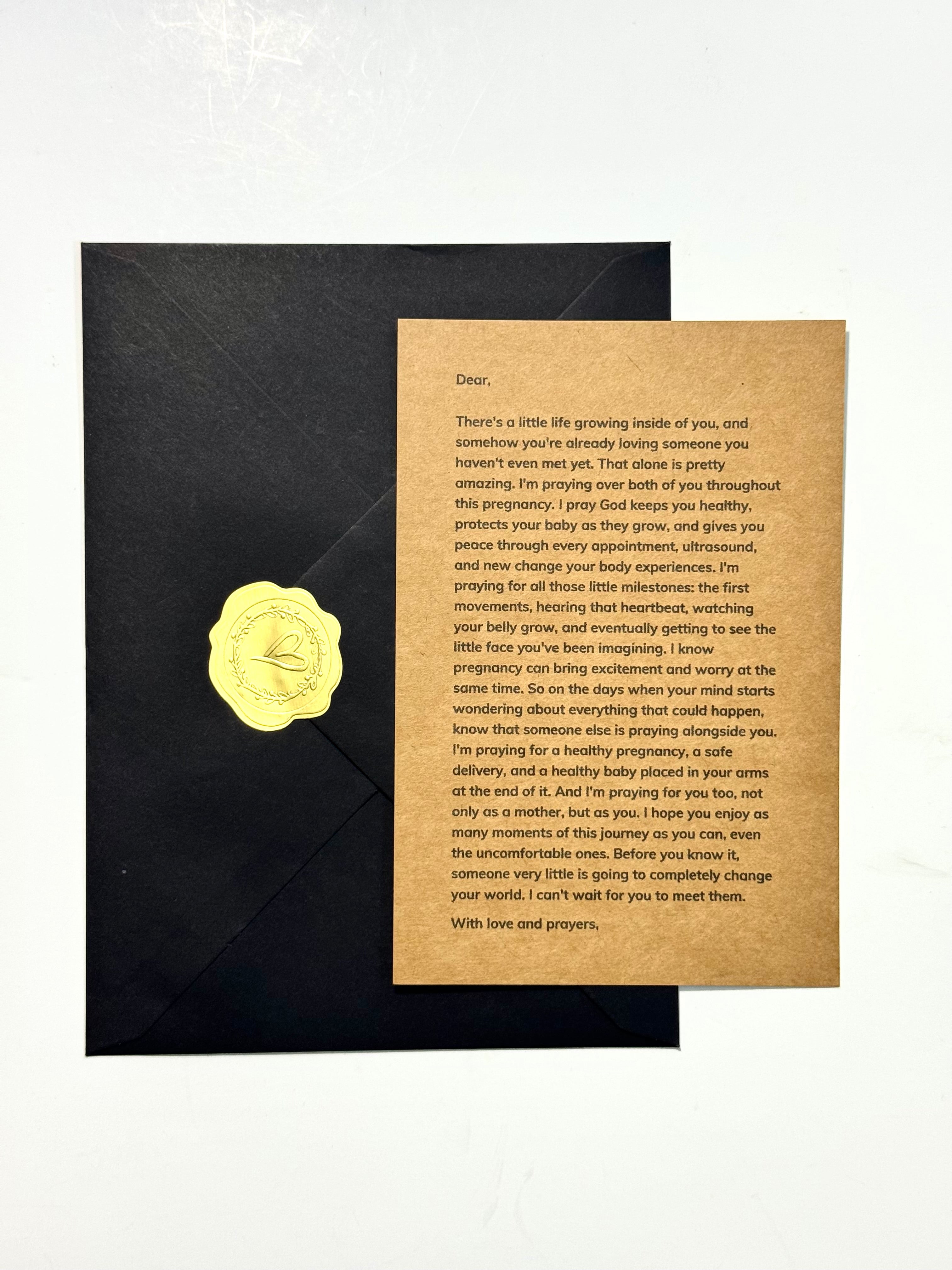A printed card with a gold embossed profile of a baby on the left, placed on top of a black envelope against a light gray background.