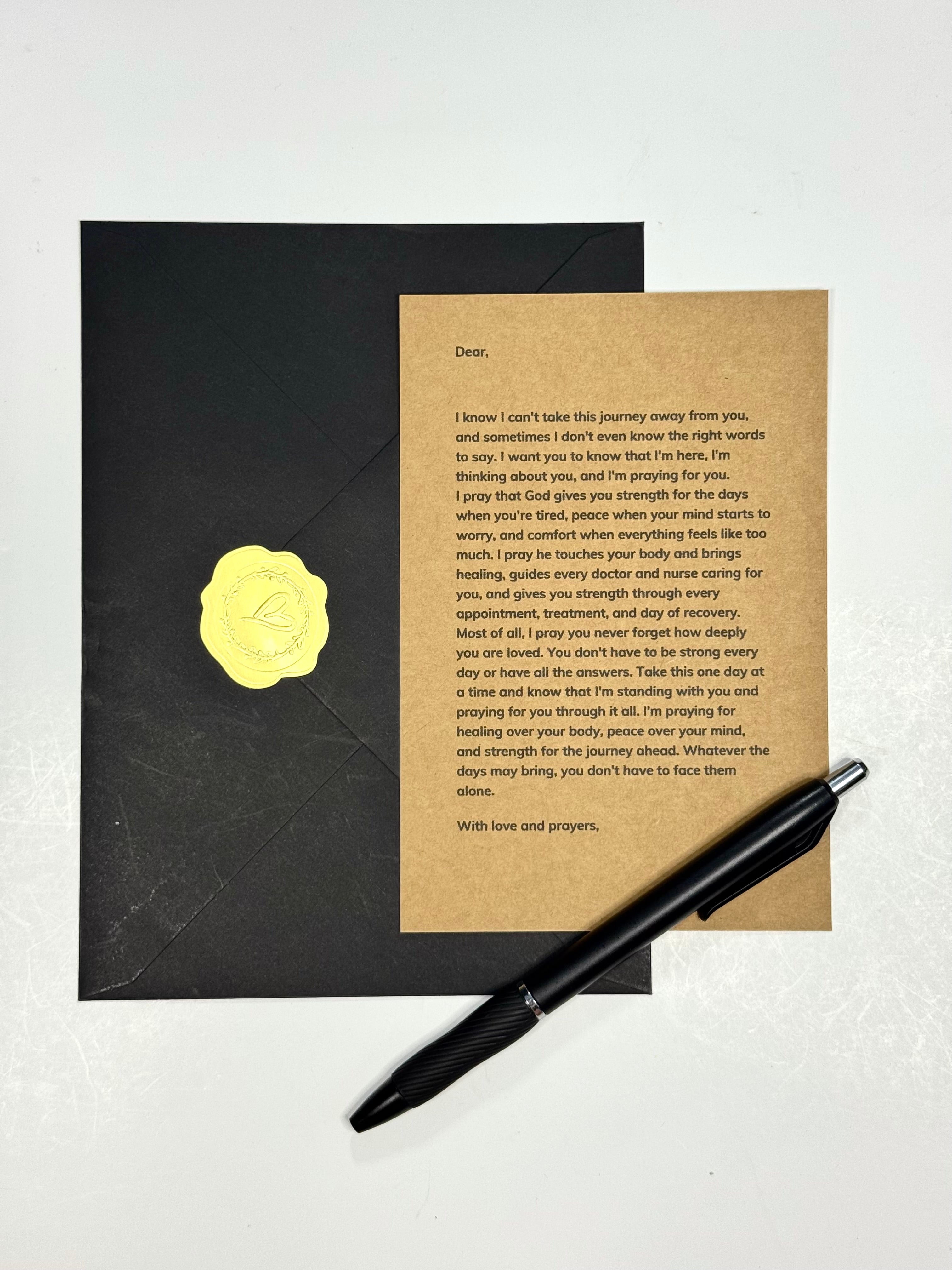 A black envelope with a gold foil seal lies next to a printed letter on a light beige card, with a black pen placed in front, all arranged on a white surface.