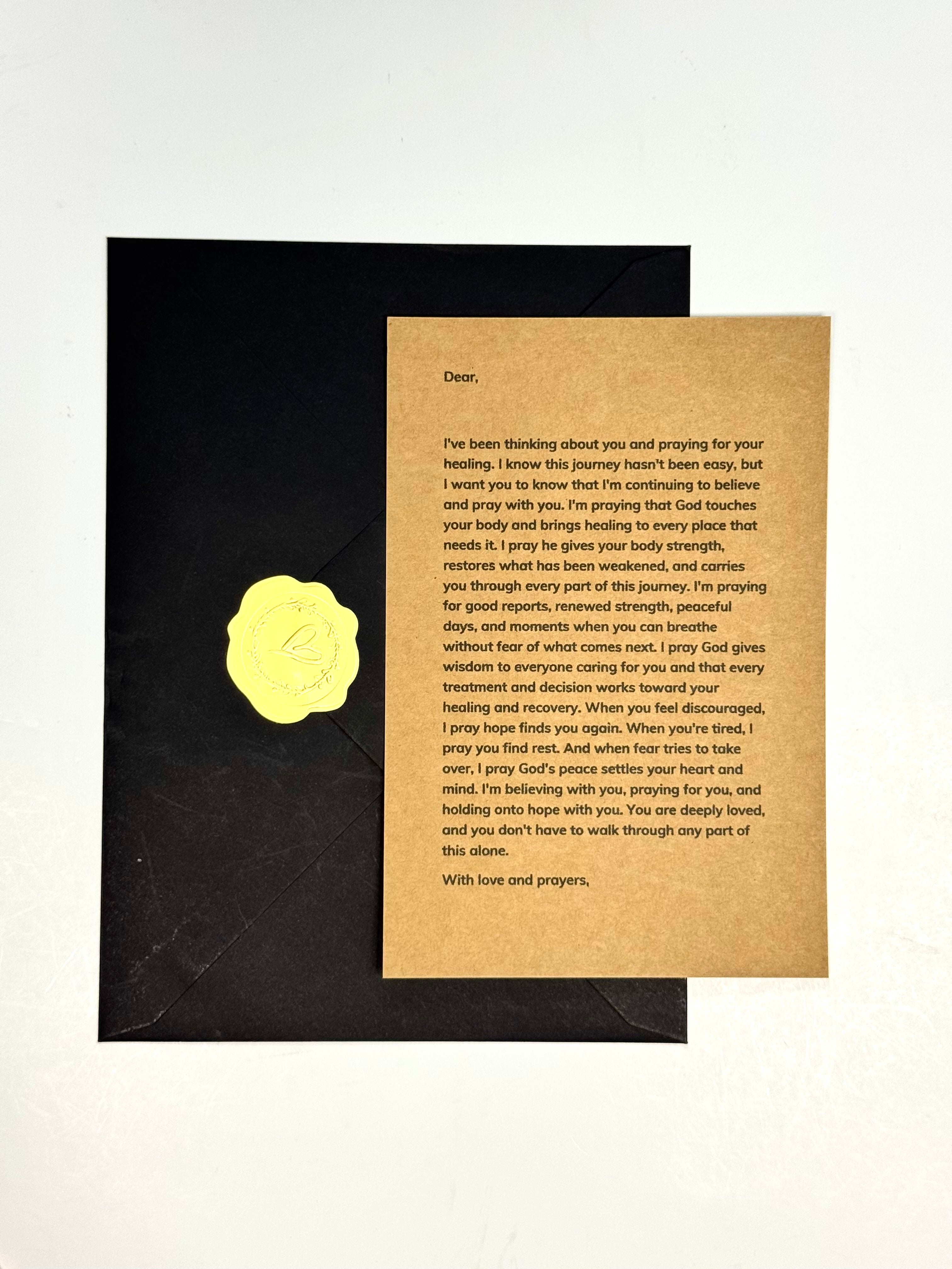 A handwritten prayer card on textured kraft paper, placed on top of a black envelope against a white background.