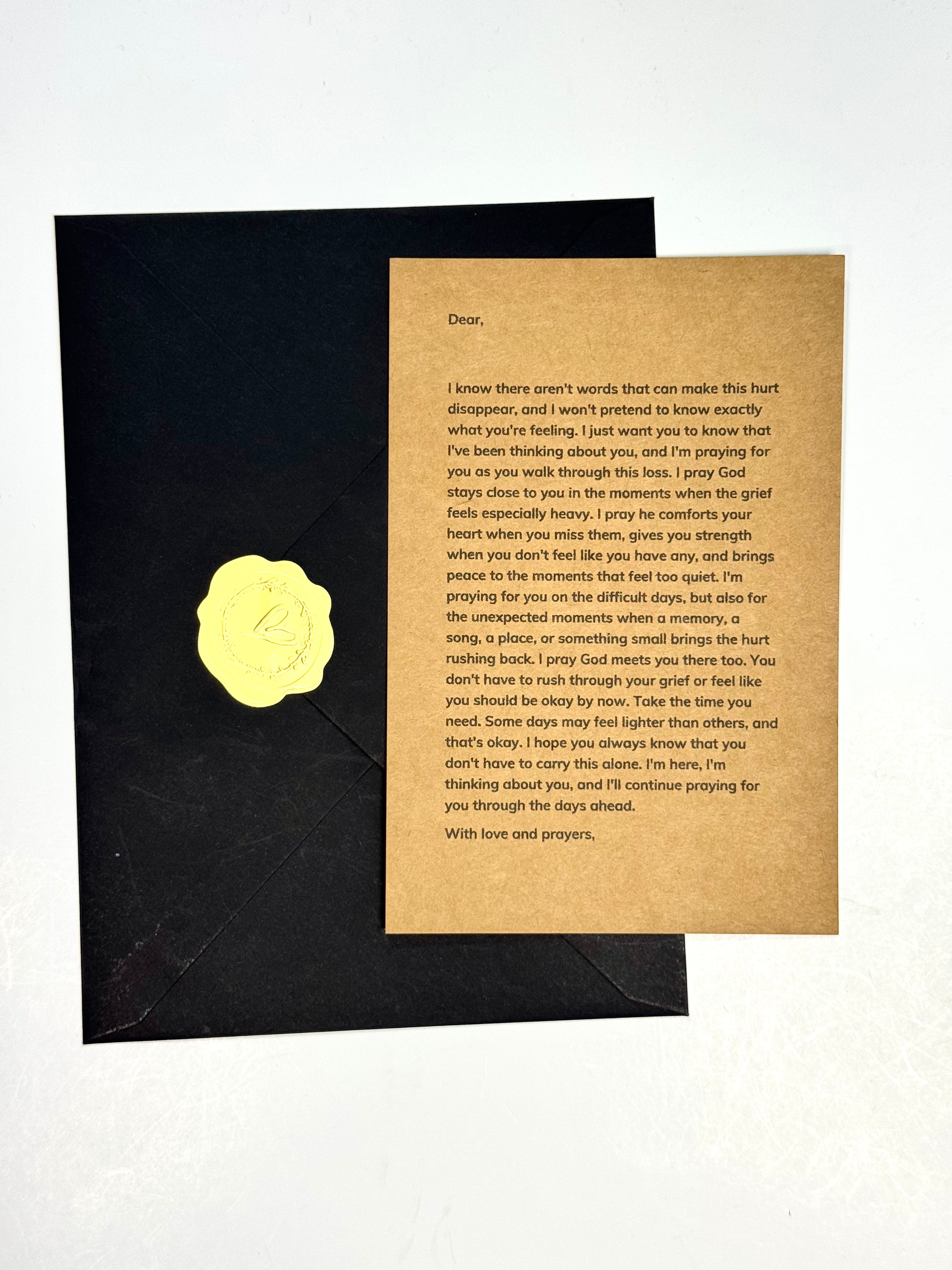 A square kraft paper condolence card with printed sympathy message and a gold foil heart, shown next to a black envelope on a white background.
