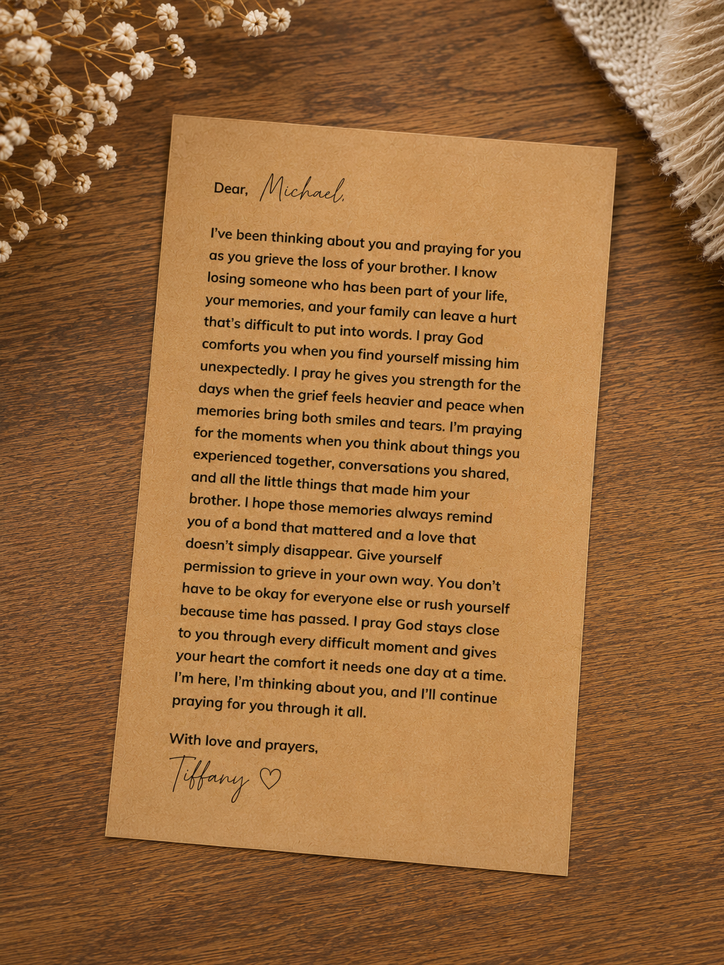 A handwritten condolence letter on a tan parchment-style card, placed on a brown wood surface with a small white floral arrangement in the background.