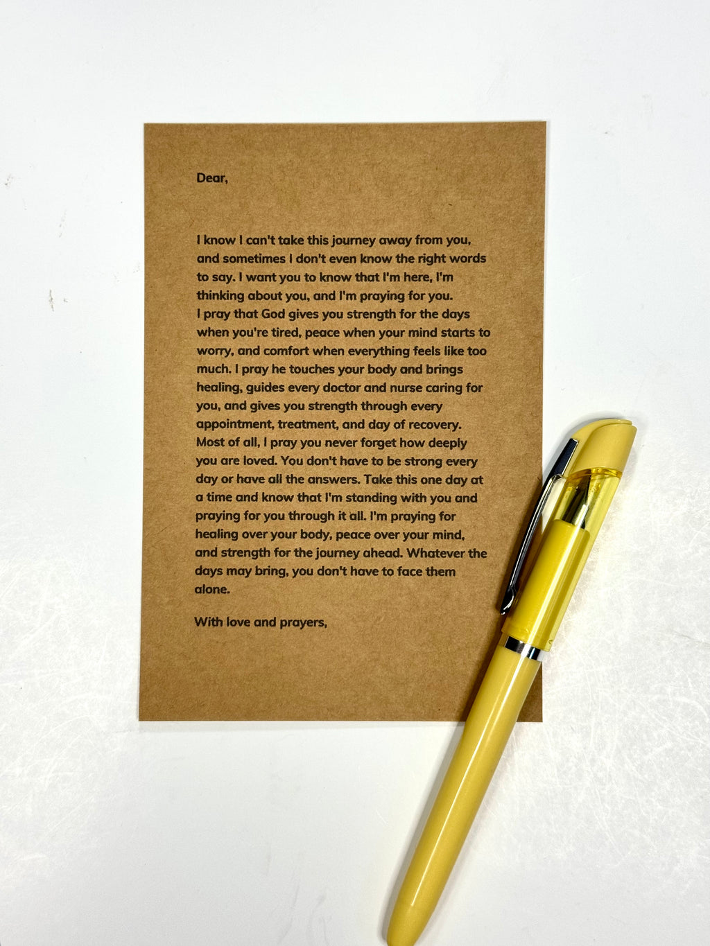 A brown kraft paper card with a handwritten letter printed in black ink, placed on a white textured background next to a gold pen.