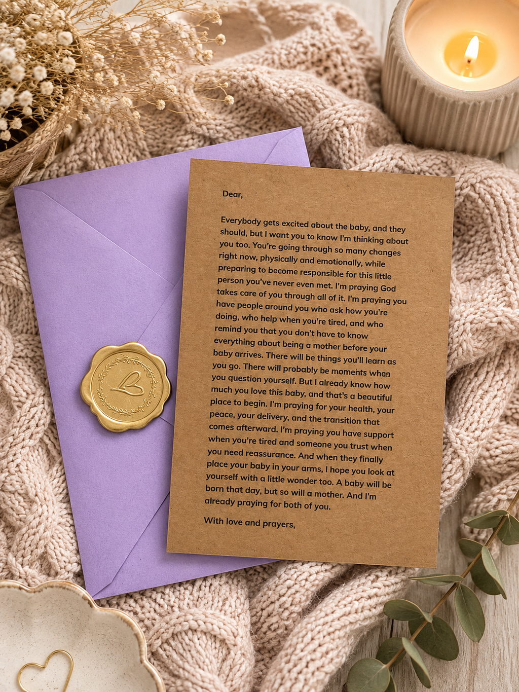 A printed baby shower congratulations card with a brown kraft-paper style background and gold foil heart details, layered over a purple envelope. The card is styled in a cozy flat-lay arrangement with a lit candle, beige knit blanket, dried flowers, and gold heart-shaped ornaments.