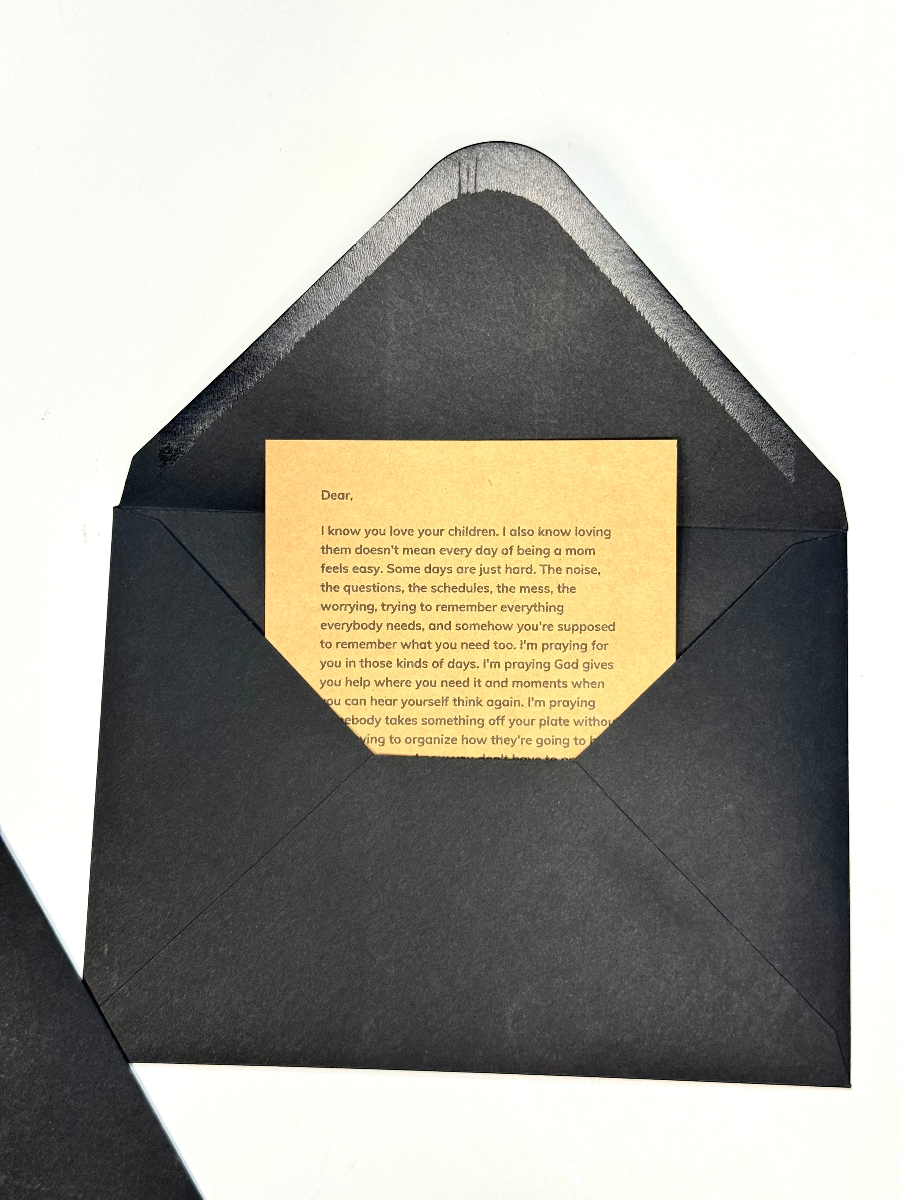 A black envelope with a blank beige card inside, shown on a plain white background. A second matching black envelope is partially visible in the lower-left corner.