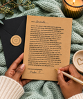 A handwritten sympathy card on brown kraft paper is held over a black fabric surface by two hands, with a gold candle in a glass holder and greenery in the background.
