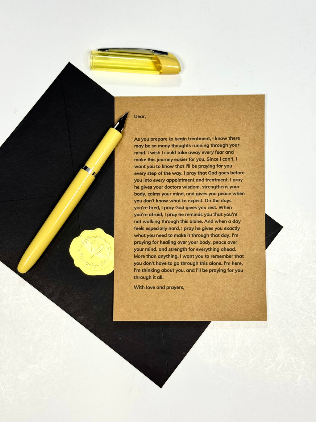 A brown kraft paper prayer card with printed prayer text, laid on a black graduation cap with a gold diploma pen and a gold pen placed beside it on a white background.