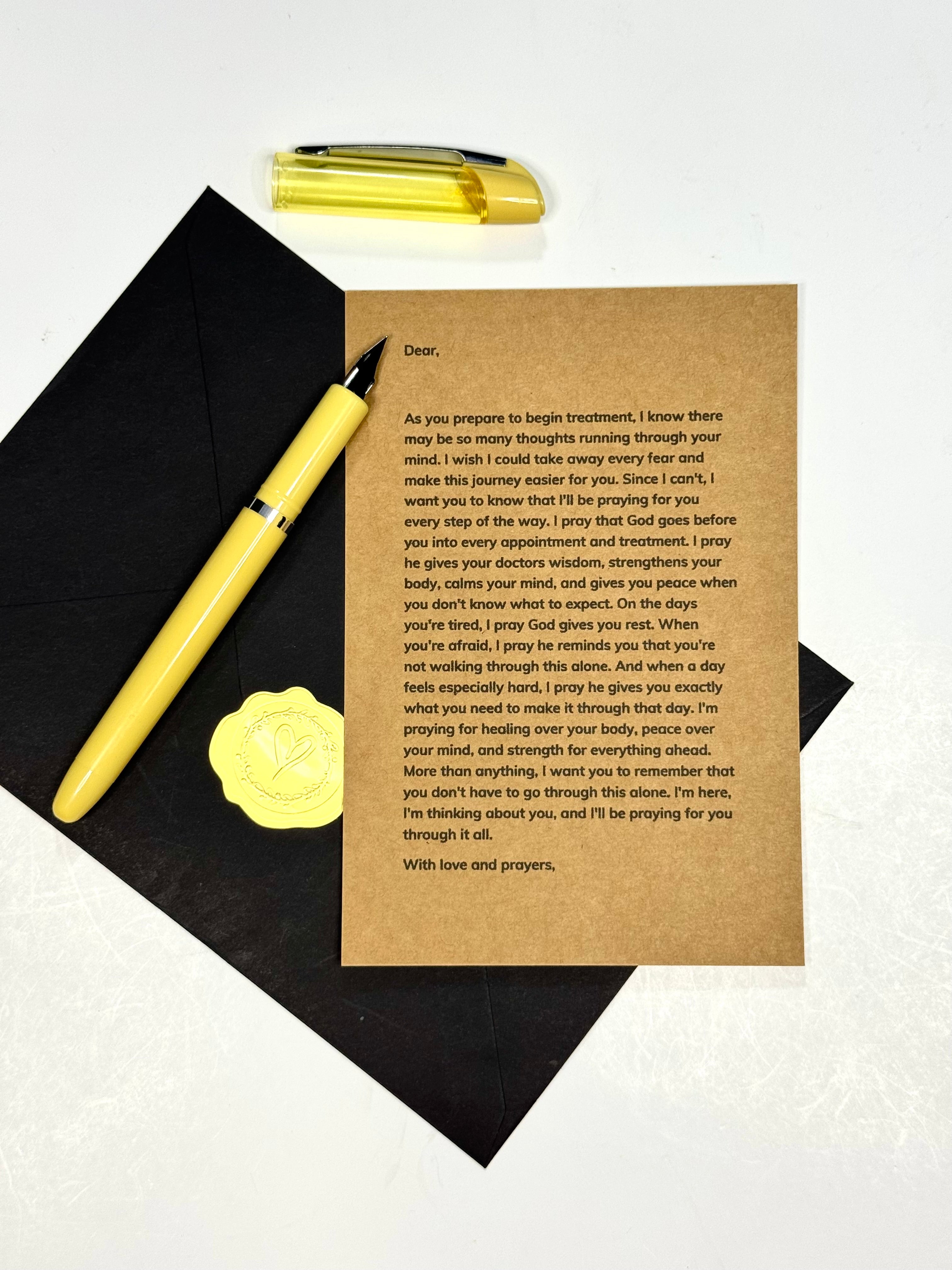 A brown kraft paper prayer card with printed prayer text, laid on a black graduation cap with a gold diploma pen and a gold pen placed beside it on a white background.