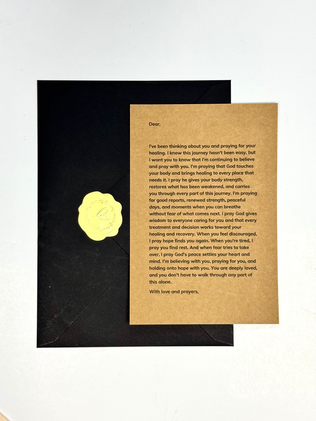 A black envelope with a gold foil seal lies next to a folded kraft paper prayer card with printed text, photographed on a plain white background.