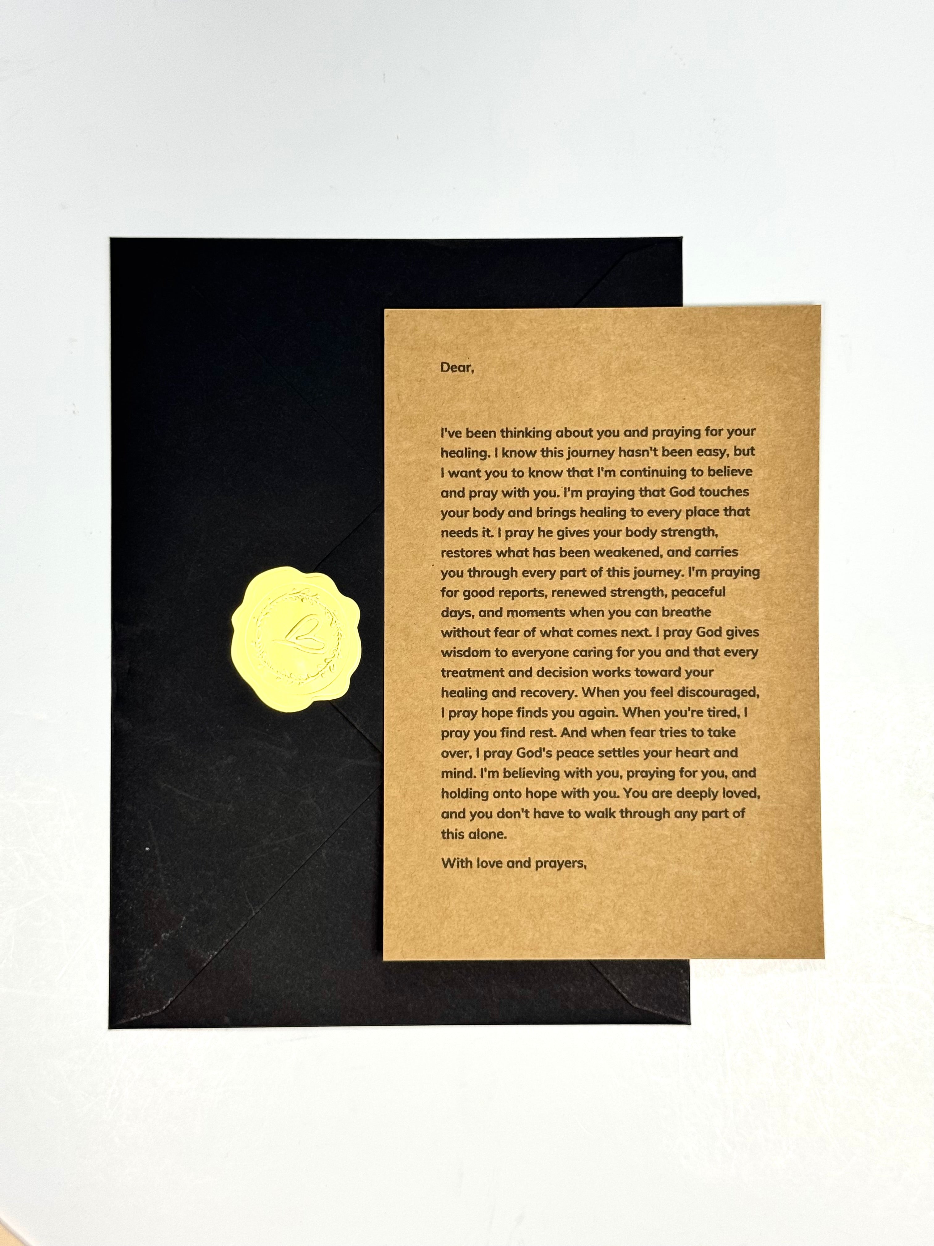 A black envelope with a gold foil seal lies next to a folded kraft paper prayer card with printed text, photographed on a plain white background.