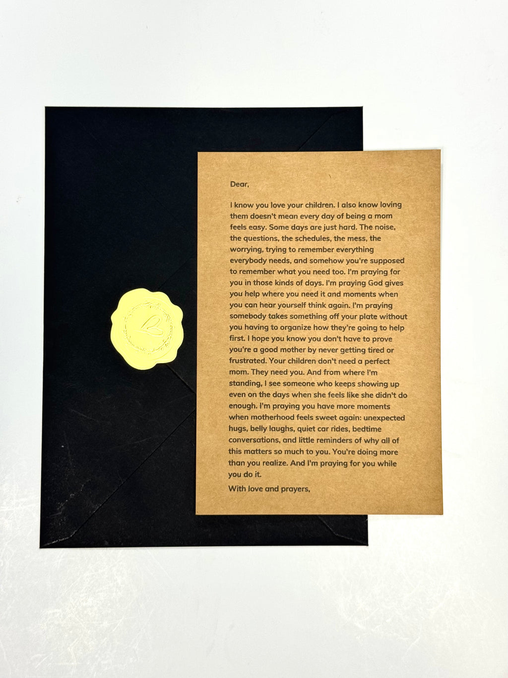 A printed apology letter card with black text on a tan background, placed on top of a black envelope with a gold wax seal, photographed on a light neutral surface.