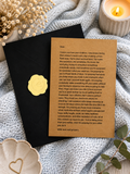 A printed prayer card for a mother's remembrance, placed on a black notebook with a gold foil seal, surrounded by dried flowers, a lit candle, and a knitted blanket.