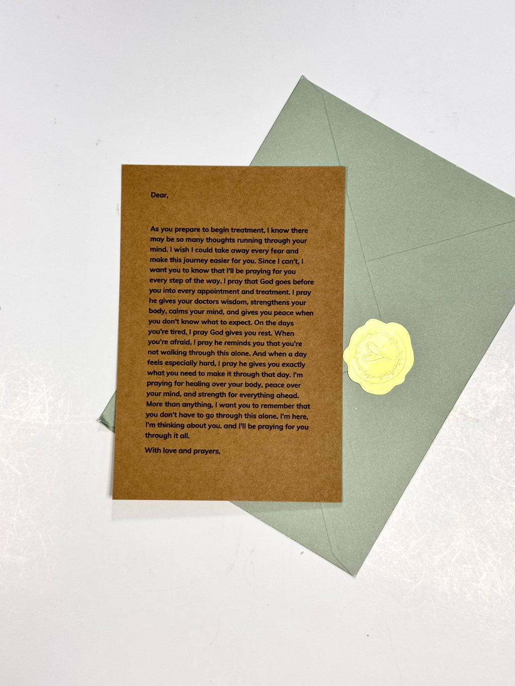 A brown kraft paper prayer card with black printed text, placed on top of a light sage green envelope with a yellow wax seal, photographed on a white background.
