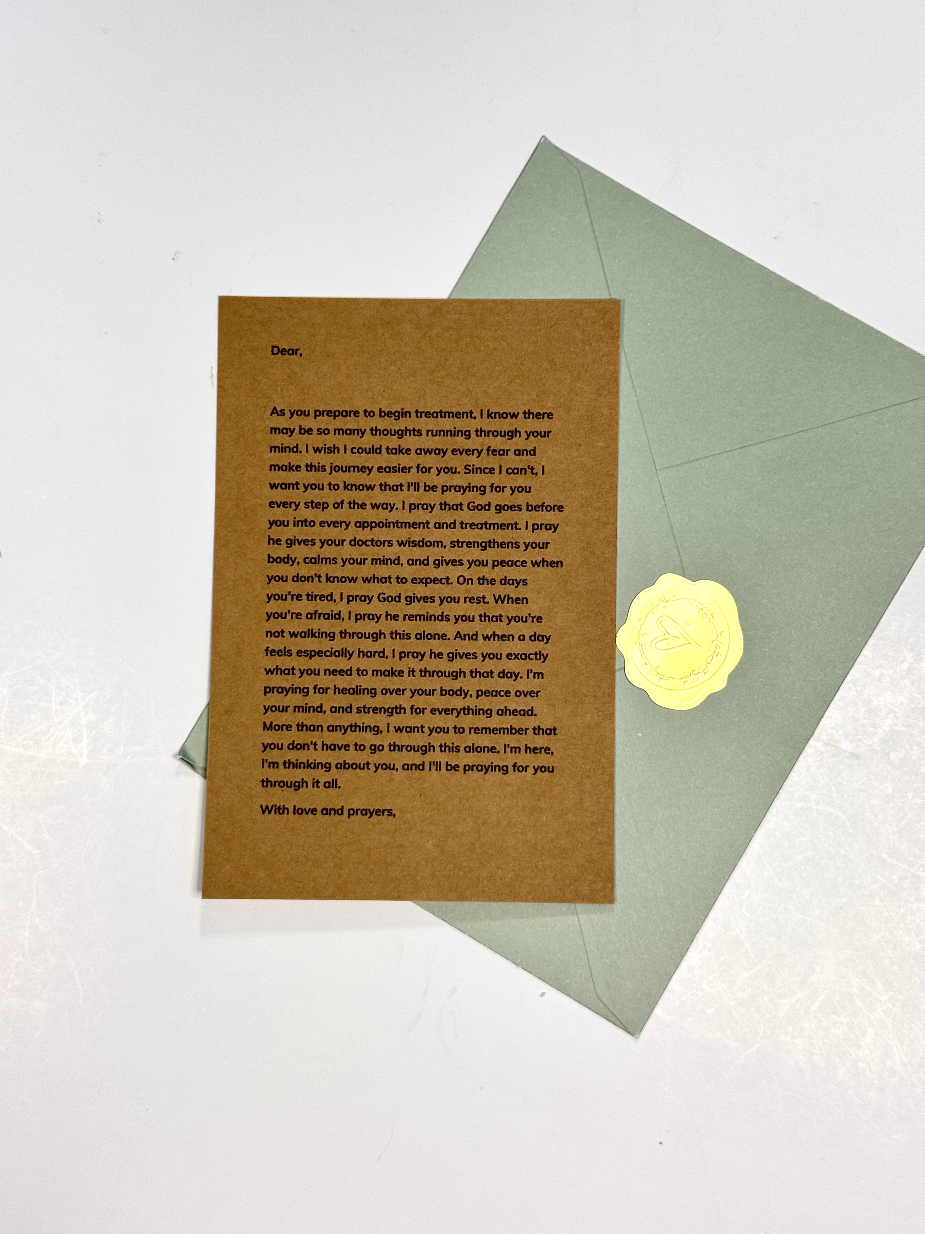 A brown kraft paper prayer card with black printed text, placed on top of a light sage green envelope with a yellow wax seal, photographed on a white background.