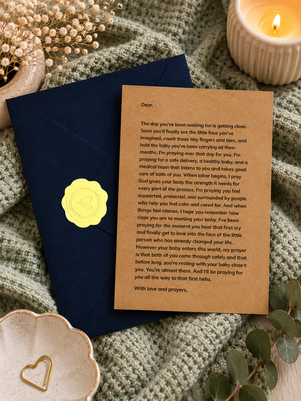 A brown kraft paper birth story card with black text, sealed with a yellow wax seal, resting on a navy blue envelope surrounded by dried flowers, a lit candle, and gold-toned decorative pieces.
