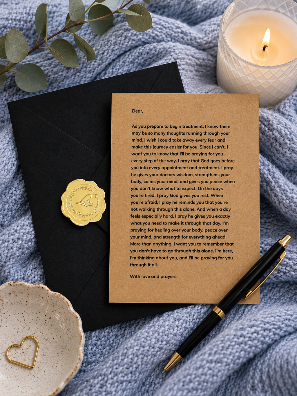 A printed prayer card with a gold embossed seal, styled in a flat-lay with a black envelope, a lit candle, a gold heart-shaped dish, and a blue textured fabric background.