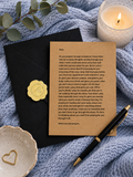 A printed prayer card with a gold embossed seal, styled in a flat-lay with a black envelope, a lit candle, a gold heart-shaped dish, and a blue textured fabric background.