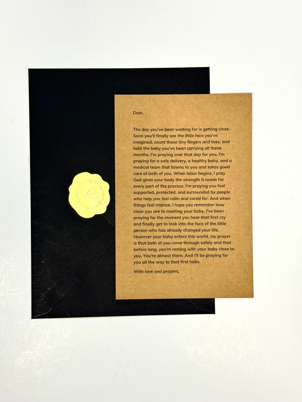 A square kraft paper prayer card with printed text, placed on top of a black envelope with a gold foil seal, photographed against a light neutral background.