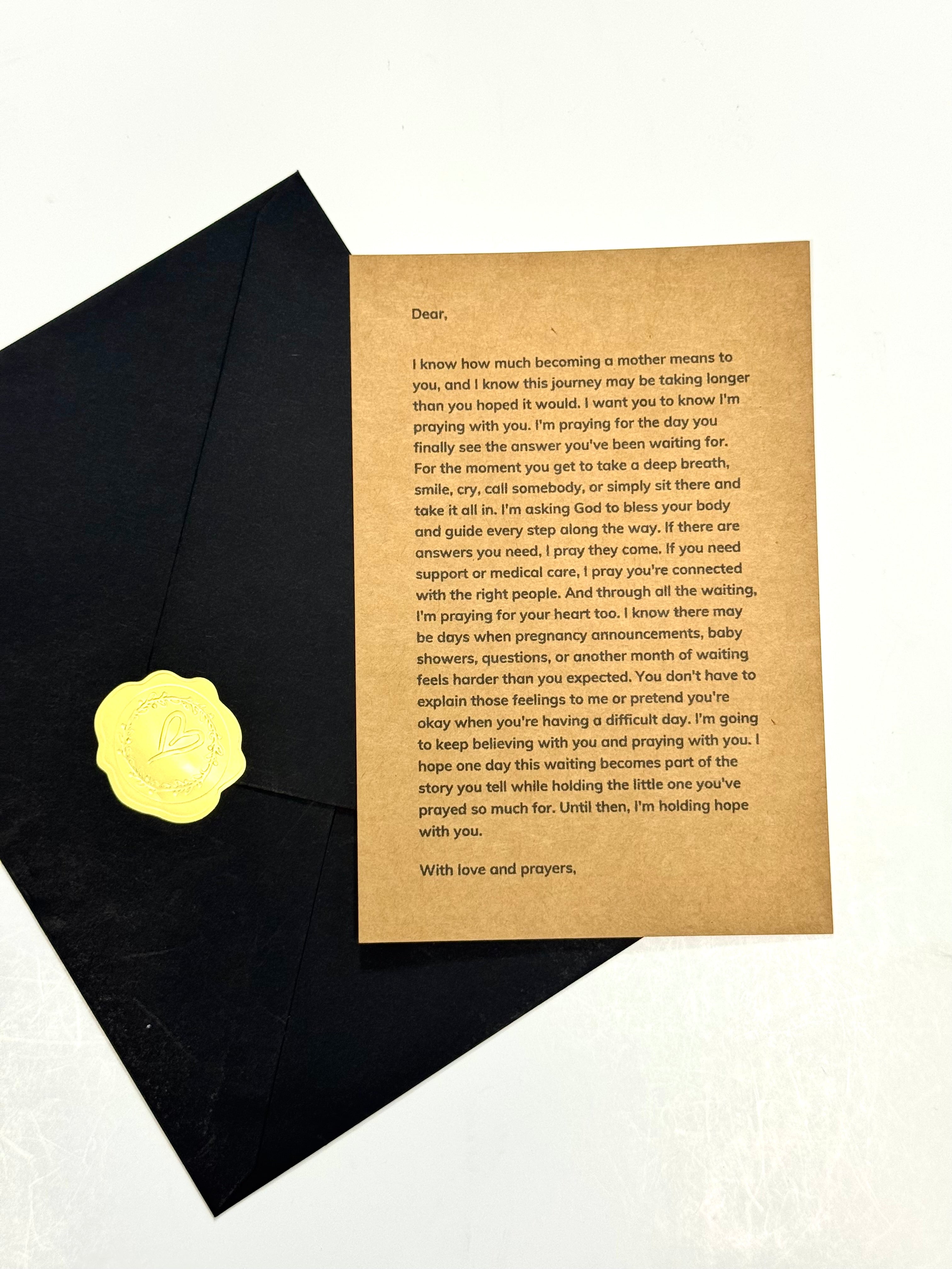 A printed letter card with black text on a tan background, placed on top of a black envelope with a gold circular seal, photographed on a light neutral surface.