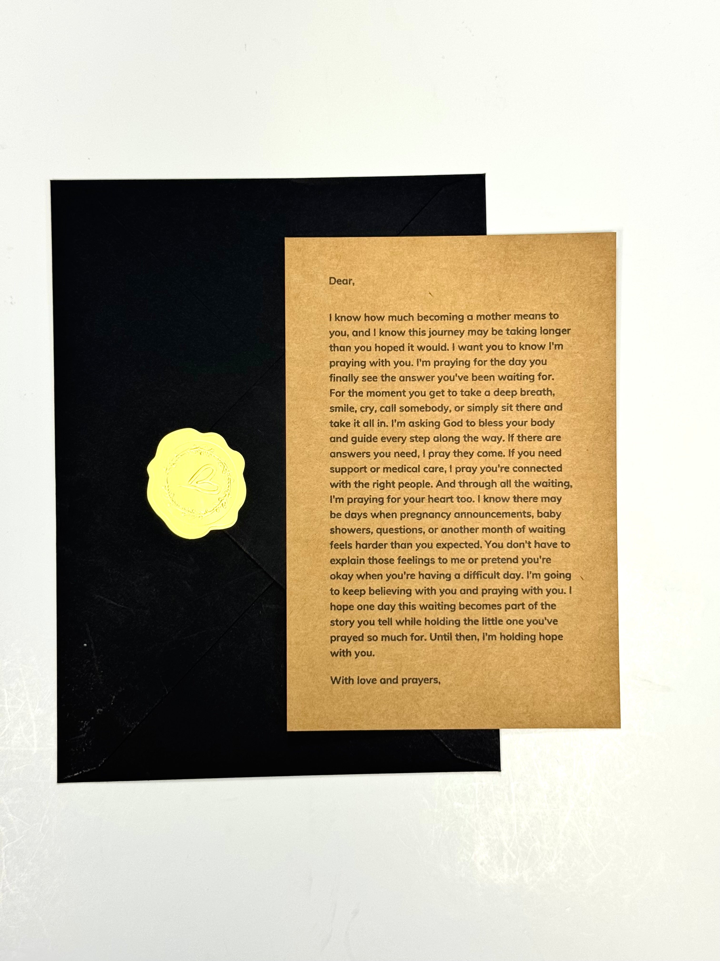 A printed prayer card with black text on a tan background, placed on top of a black envelope against a white backdrop.
