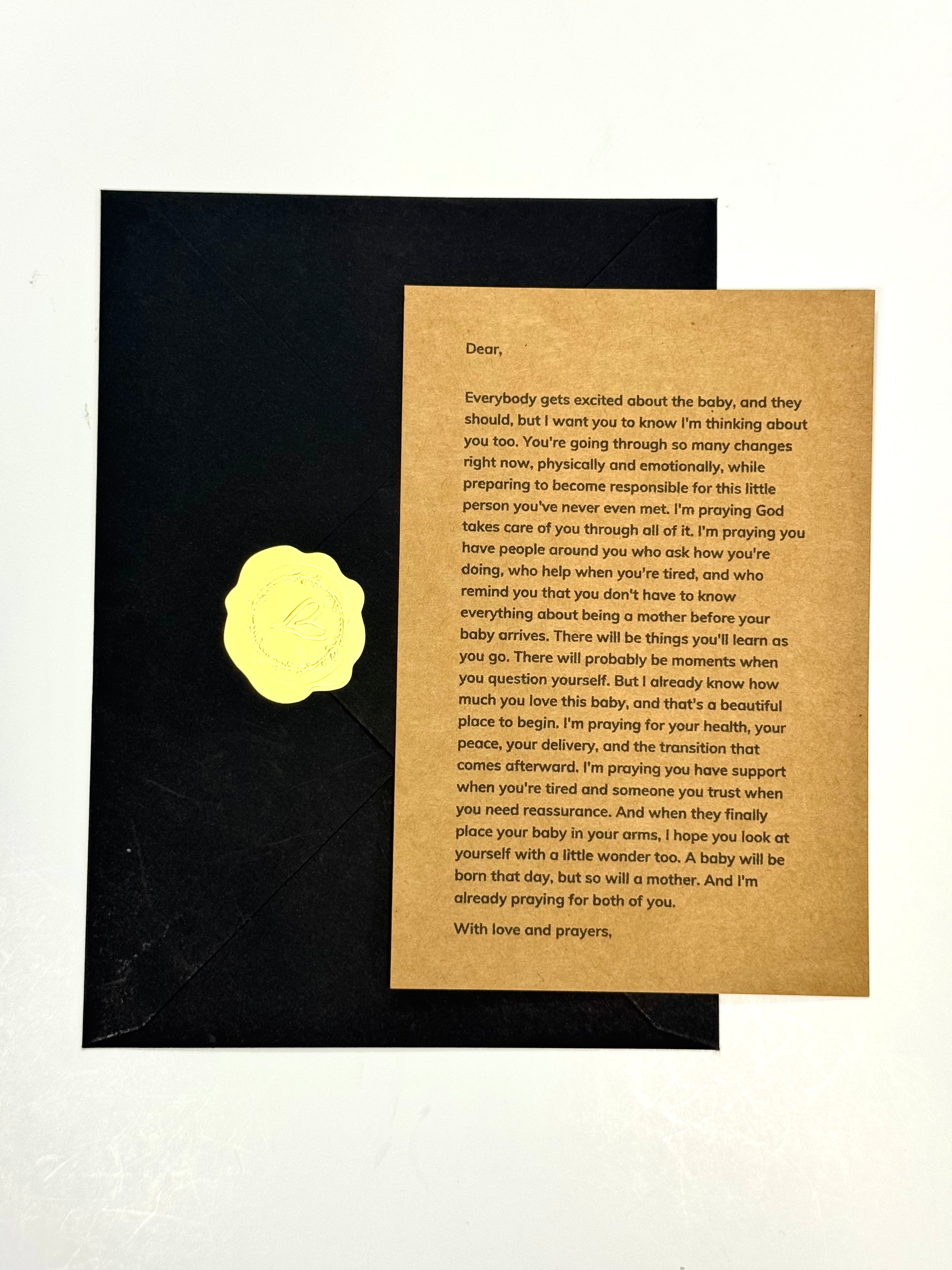 A printed prayer card with black text on a tan background, placed on top of a black envelope against a white backdrop.